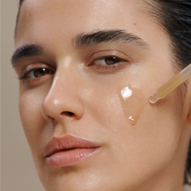 Close-up of a woman's face with a clear gel dropper applying a transparent serum to her cheek against a soft beige background.