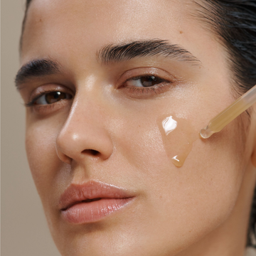 Close-up of a woman's face with a clear gel dropper applying a transparent serum to her cheek against a soft beige background.