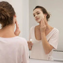 Someone applying a white facial mask or skincare product while looking in a bathroom mirror.