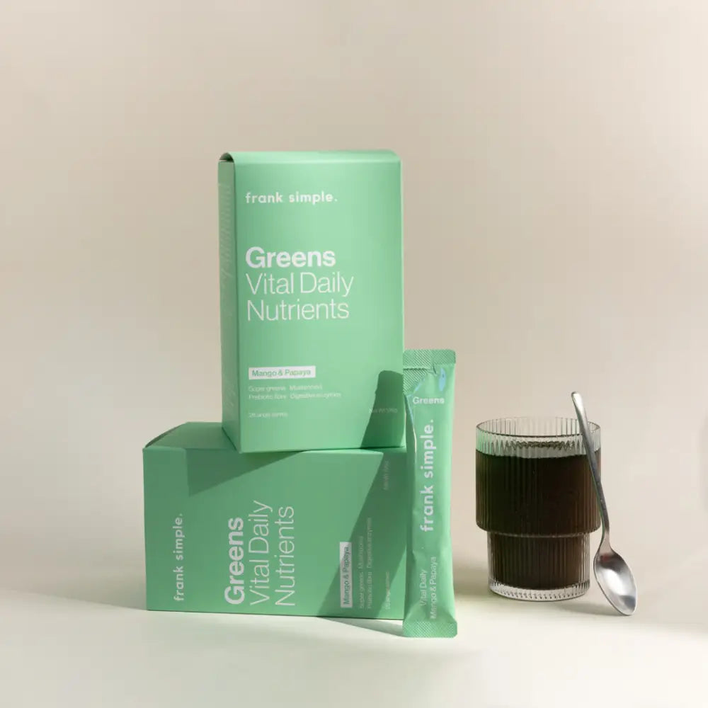 Mint green supplement boxes labeled ’Greens Vital Daily Nutrients’ with a glass of dark liquid beside them.