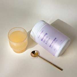 Purple and white cylindrical container of Morning Glory collagen powder alongside a glass of liquid and gold spoon.