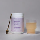 White cylindrical container labeled ’Morning Get Up & Glow’ with a golden spoon and glass of pale liquid beside it.