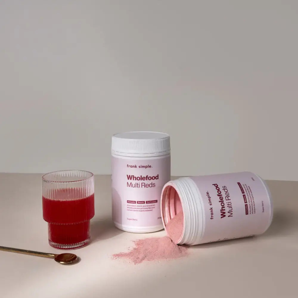 Pink nutritional supplement powder spilling from a white container alongside a glass of red liquid.