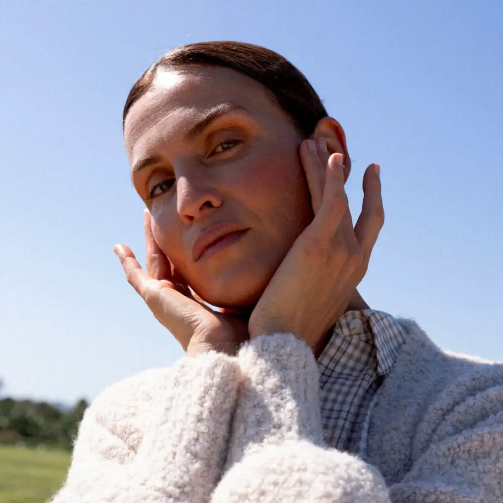 A person wearing a white fuzzy sweater and checkered shirt while touching their face.