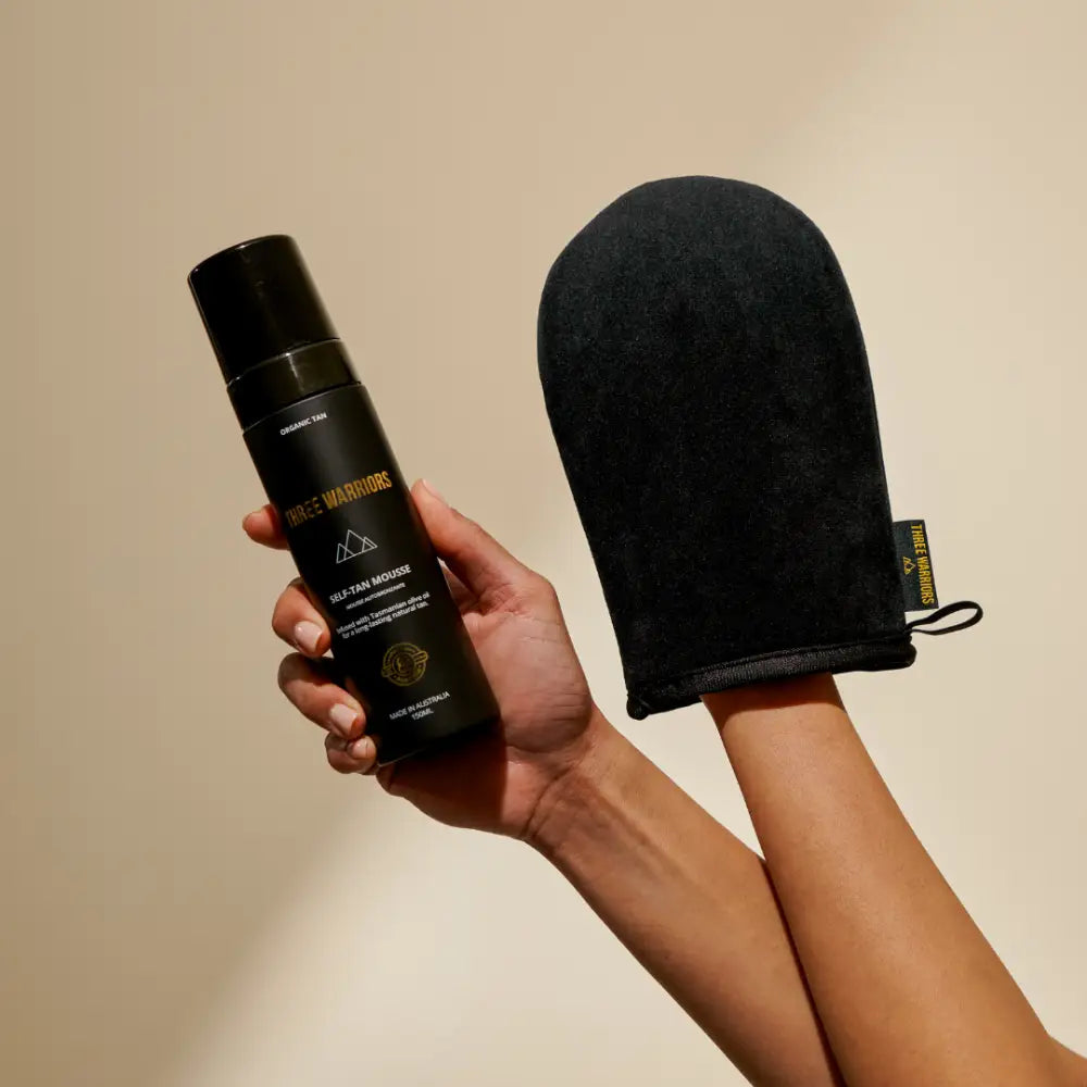 Black self-tanning mitt and tanning product bottle being held up against a neutral backdrop.